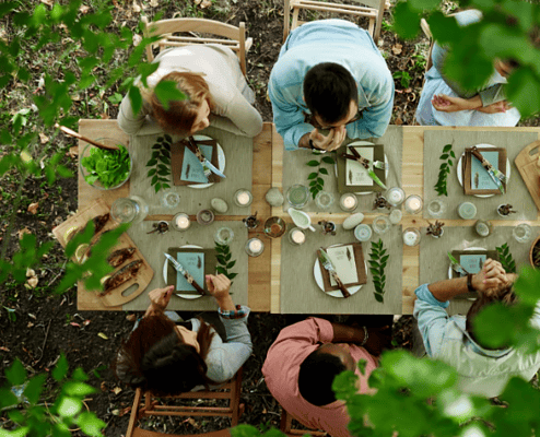 Overhead view of people seated around a long wooden table enjoying an outdoor meal surrounded by greenery, with candles, place settings, and shared dishes arranged for a communal dining experience.