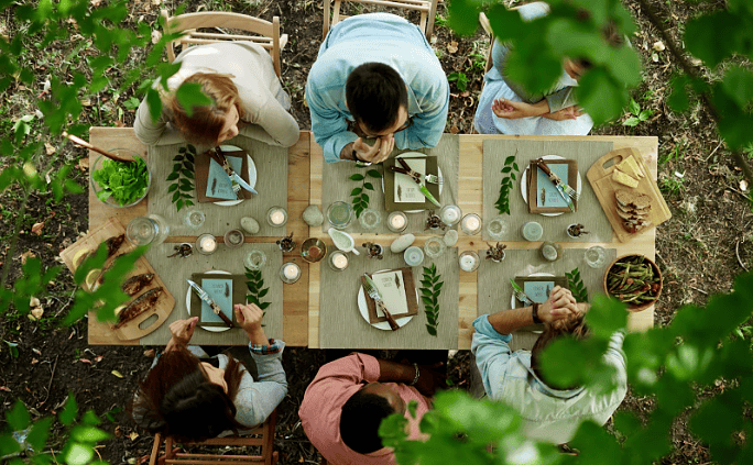 Overhead view of people seated around a long wooden table enjoying an outdoor meal surrounded by greenery, with candles, place settings, and shared dishes arranged for a communal dining experience.