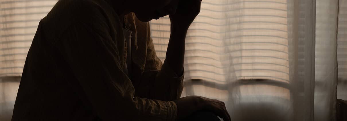 A person sitting by a window with blinds, appearing distressed or contemplative, illustrating emotional struggles related to ADHD self-blame.