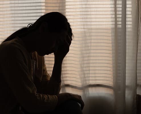 A person sitting by a window with blinds, appearing distressed or contemplative, illustrating emotional struggles related to ADHD self-blame.
